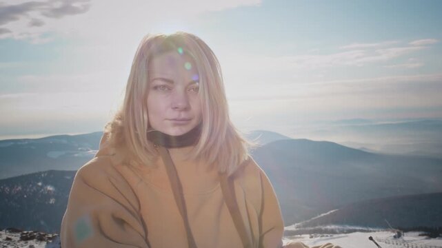 Portrait of a lovely girl enjoying ski season vacations. Blond hair moving in the wind in slow mo. Dressed in a hoodie, she fixes her hair with her hand. Mountain peaks in the snow. Winter travel.