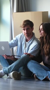 Young happy couple sitting on the floor of their new apartment among cardboard boxes, using a laptop to plan renovations and shop for furniture and decorations online together
