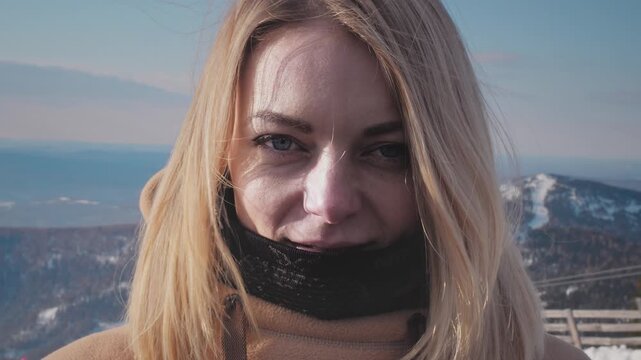 Portrait of a lovely girl enjoying ski season vacations. Blond hair moving in the wind in slow mo. Dressed in a hoodie, she fixes her hair with her hand. Mountain peaks in the snow. Winter travel.