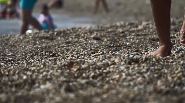 Close up view of person bare feet walking slowly on coarse pebble beach. Sunlight glinting on wet stones. Blurred background people enjoying shallow ocean water. Summer day activity. Low angle