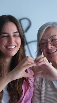 Happy young woman and her elderly grandmother looking at the camera, smiling, and making a heart shape with their hands together, symbolizing their strong intergenerational family bond and love