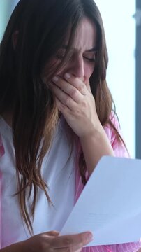 Upset young woman reading a document with terrible news, bursting into tears and covering her mouth in shock and despair while standing near a window at home or in an office