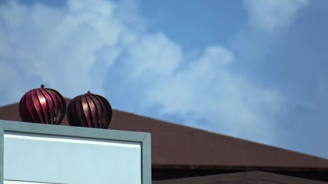 Close up view of two reddish-bronze metallic spherical objects with ribbed surface on top of building roof. Expansive blue sky with scattered white clouds above. Bright sunny day, architectural detail