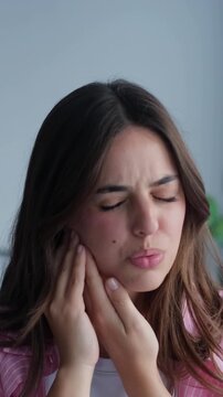 Unhappy young woman touching her cheek and grimacing from a sudden toothache, suffering from severe dental pain, sensitive teeth, caries, or pulpitis problems at home, needing a dentist