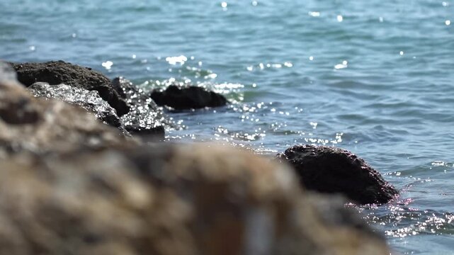 Close-up view of sunlit ocean water with gentle waves lapping against rough, dark rocks on a sunny day. Surface reflects bright sunlight, creating shimmering effect as water recedes, revealing wet tex