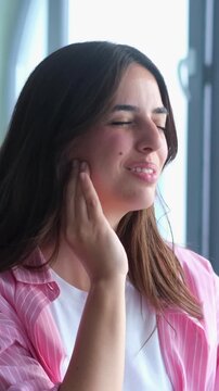 Unhappy young woman with long hair suffering from a severe toothache, touching her cheek with her hand and showing an expression of intense pain due to dental problems and sensitivity
