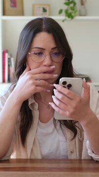 Beautiful thoughtful young woman wearing glasses sitting at a table at home, scrolling and using her smartphone for communication, showing various emotions like surprise, concern, and boredom