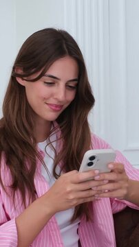 Beautiful smiling caucasian woman using her mobile phone for texting, browsing social media, or online shopping while relaxing on the couch at home, enjoying a pleasant conversation