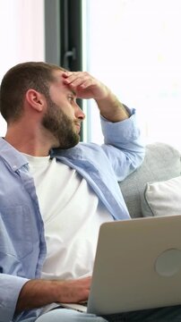 Worried, tired man feeling headache and stress from remote work, rubbing his forehead and hair while sitting on the sofa with a laptop, looking thoughtful and concerned about a problem
