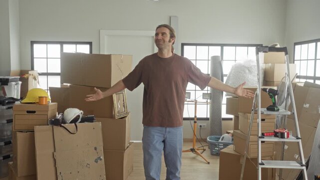 Man with outstretched arms and visible palms amid stacked cardboard boxes, ladder and cordless drill in a sunlit room inside a building; hopeful new beginning.