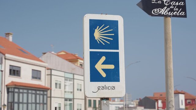 Camino de Santiago waymarker sign featuring a yellow scallop shell and left arrow on a blue background, located in a town in Galicia.