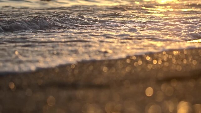 Close up view of small ocean waves washing onto sand shore, illuminated by golden light of sunset. Sunlight creates shimmering bokeh effect on beach foreground, highlighting peaceful movement of water
