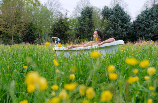 Woman relaxing in a vintage bathtub placed in a blooming meadow surrounded by tall grass and yellow flowers creating a surreal outdoor nature scene