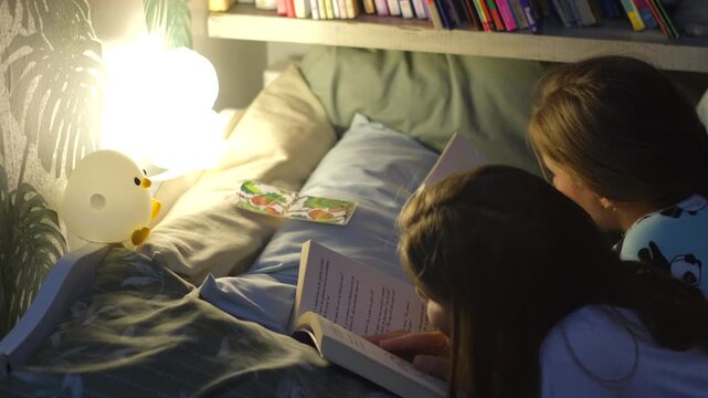 Twin sisters reading bedtime stories together in single bed near nightlight