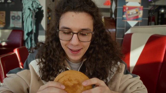 Medium close-up of nerdy young Caucasian man sitting at diner, rubbing hands, biting at large hot chili cheeseburger, experiencing intense burning sensation in mouth and drinking soda to extinguish it