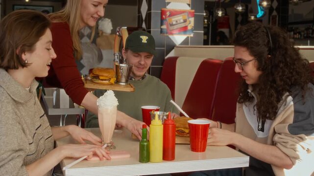 Medium shot of young Caucasian fast food restaurant waitress bringing milkshake, two portions of appetizing hamburgers and fries for group of young gen Z students chatting and laughing at table