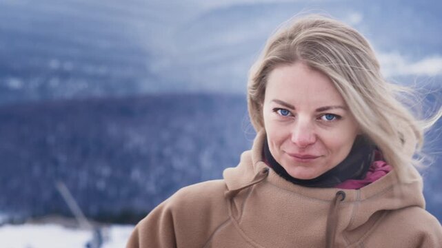 Portrait of a beautiful girl will fix her hair in winter. Beautiful big blue Eyes, Wind blowing on Blonde Hair. Happy skiing. Dressed in a hoodie. Travel to winter resort, adventure. Slow motion