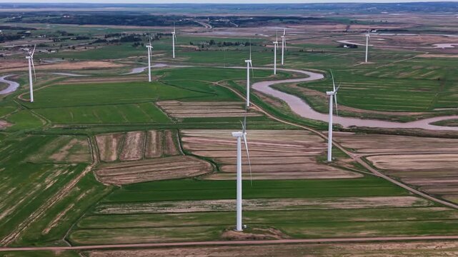 Cinematic drone shot aerial view of a field in Nova Scotia, Canada. Windmills stand in rows near a river and the ocean. The landscape features green fields and water