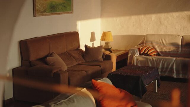 Warm, sunlit interior of a cozy living room at golden hour, featuring comfortable brown sofas and a lamp casting a soft glow, creating an atmosphere of peaceful relaxation and domestic tranquility.