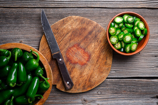 Cutting board with knife and bowl of jalapeno slices on wooden surface. Fresh green chili preparation with seeds visible