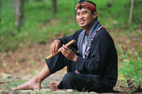 An Indonesian Sundanese man wearing traditional black Pangsi clothing and batik Iket headpiece, holding a bamboo flute in a natural outdoor park.