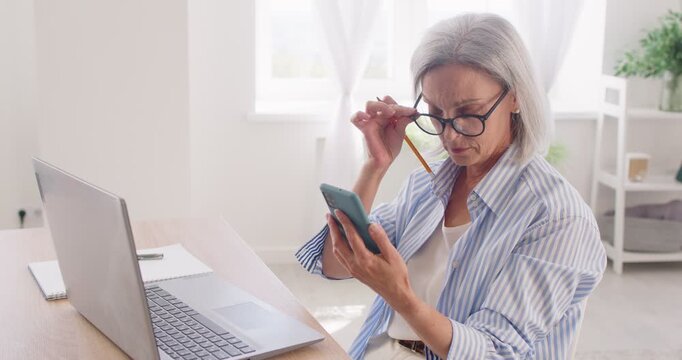 Focused mature woman in eyeglasses sitting at desk in front of laptop, looking through information on mobile phone. Outraged nervous busy senior gray haired lady having problems with work issues.