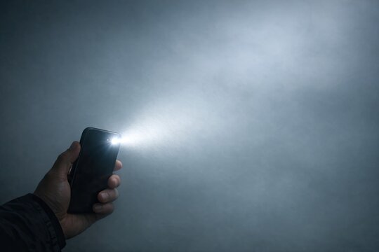 A person holds a cell phone with a flashlight shining on it, suitable for use in scenes where a character is trying to find their way or reading something