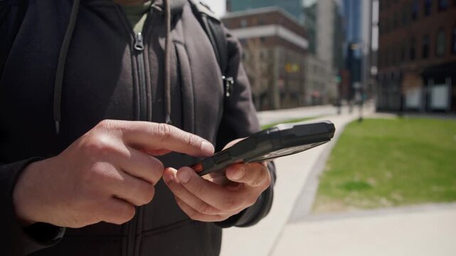 A man stands on a lively street, interacting with his smartphone. The close-up showcases his hand as he navigates the gadget, connecting with the digital world around him