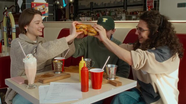 Medium slowmo shot of young Caucasian gen Z girl and two guys enjoying gourmet burgers at retro diner after successful college exams, toasting each other and cheering with happy smiles