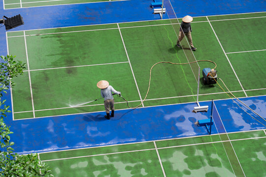 Workers clean wet tennis courts using hoses and spraying equipment at an outdoor recreational sports complex in Vietnam.