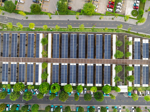 Top down drone photograph of solar panel structures installed above parking spaces along a landscaped urban roadway.