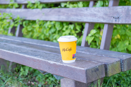 yellow McCaf&eacute; coffee cup with logo on wooden bench in park, blurred natural backdrop, modern mobility, outdoor lifestyle, fast food beverage, branded cup, ideal ads about travel, breaks consumerism