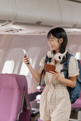 Fototapeta premium Young female passenger enjoying her flight, checking smartphone and travel documents, listening with headphones, relaxing by the window seat and expressing happiness during her air journey.