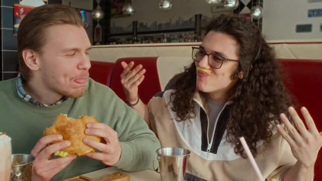 Medium portrait of young Caucasian man chewing delicious burger while sitting in diner booth, while nerdy friend in glasses joking, making horns gestures and fun mustache with fries, looking at camera