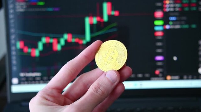 Human hand holding shiny bitcoin coin in front of laptop screen with financial trading charts