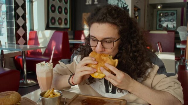 Medium portrait of young Caucasian zoomer guy with long curly hair, glasses munching at delicious burger at favorite diner while enjoying meal with milkshake and fries, looking at camera while chewing