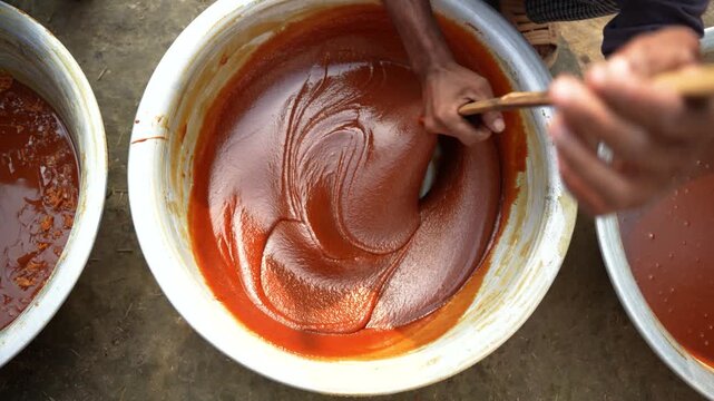 View of hands stirring thick reddish-brown traditional sweet paste in a large metal bowl with a wooden stick in Natore, Rajshahi Division, Bangladesh.