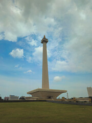 Obraz premium National monument in jakarta, a tall stone pillar with a golden flame against a blue cloudy sky, grass surroundings. Suitable for travel guides, architecture themes, and historical websites.