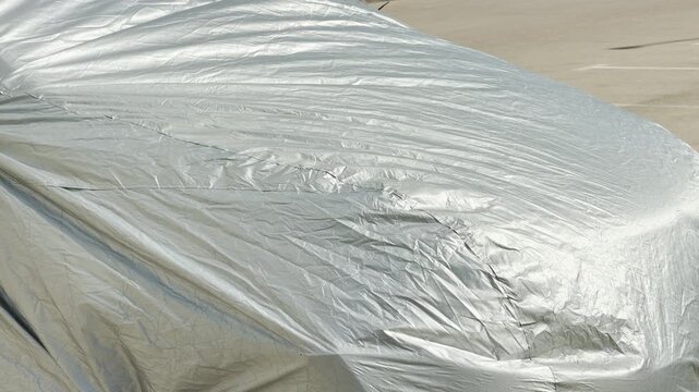 A wrinkled silver tarp tightly covers the front hood of a car. The hidden vehicle rests outside on a sunny day in an asphalt parking lot area.