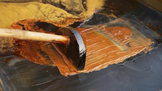 View of a wooden tool spreading thick brown molasses on a flat surface during traditional jaggery production in Natore, Rajshahi Division, Bangladesh.