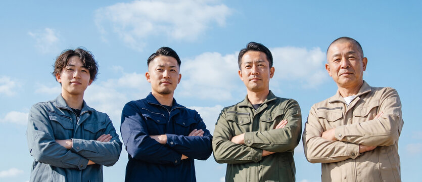 青空を背景に並んで立つ様々な年代の作業員の男性　建設現場 製造業 採用