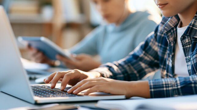 Closeup of a young student using a laptop for schoolwork soft daylight highlighting neat desk setup tutor behind holding open tablet to assist in quiet organized workspace.