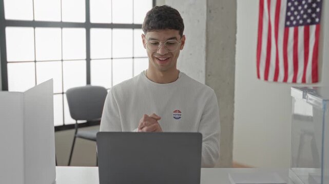 Man wearing glasses and an i voted sticker typing on a laptop with hands visible at a polling table inside a building near an american flag; civic duty pride.