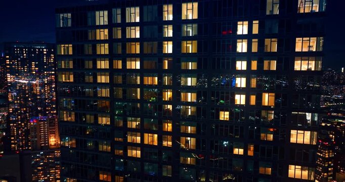 Rising along the multi-storied building with switched on illumination in the windows. City lights sparkle on the glossy facade of the high-rise at night.