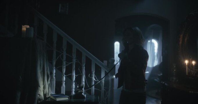 Tracking wide shot of silhouetted young woman on rotary phone as power goes out in scary home