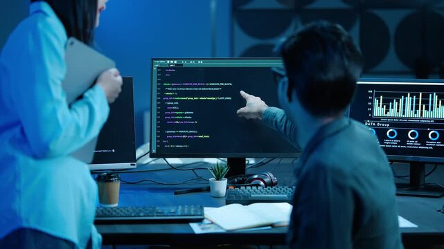 Two IT programmers collaborate on software development at night. A male coder points at the computer screen discussing code with a female colleague in a dark modern office with data dashboards.