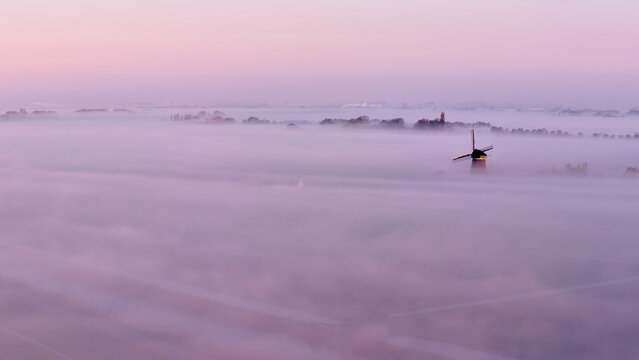 Aerial view of the Drainage mill De Vierwinden emerging through thick morning mist under a purple sunrise sky in Opmeer, North Holland, Netherlands.