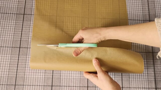 Hands holding scissors cut through brown wrapping paper, revealing a neatly trimmed sheet on a checkered surface, showcasing the progression of the cutting action