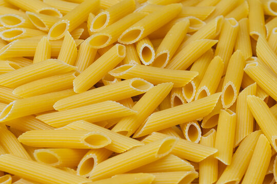 Close-up penne pasta tubes with characteristic ridges and hollow centers scattered together. Background traditional Italian pasta, for Italian cuisine, cooking ingredients and food preparation.