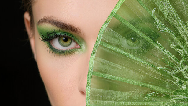 Extreme close up , A green fan is covering half face of beautiful woman , green eyes , black background.